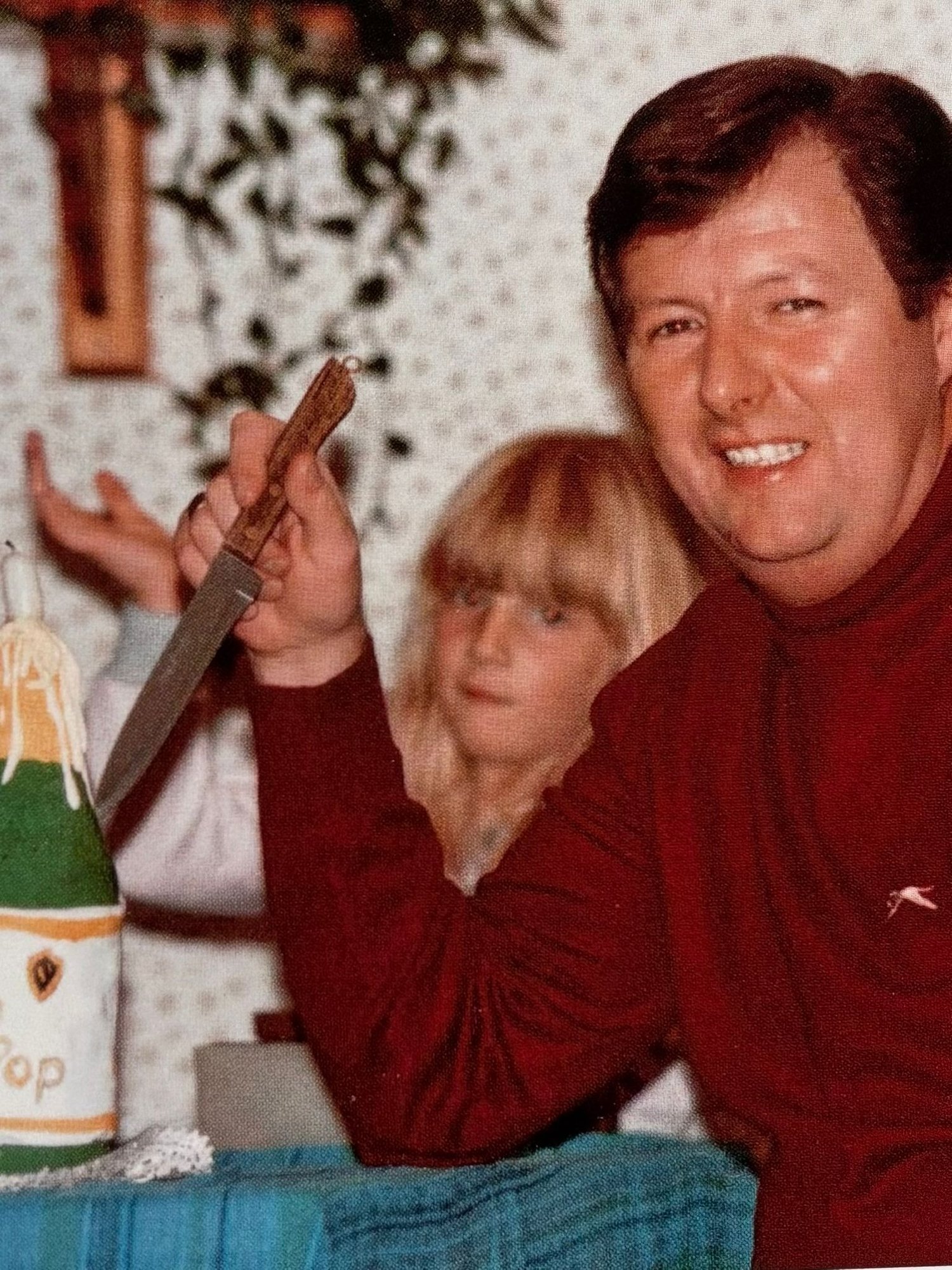 A smiling older man in a red sweater sits at a table, holding a knife poised to cut a cake shaped like a champagne bottle. The moment feels warm and celebratory, capturing his quiet presence and gentle humour.